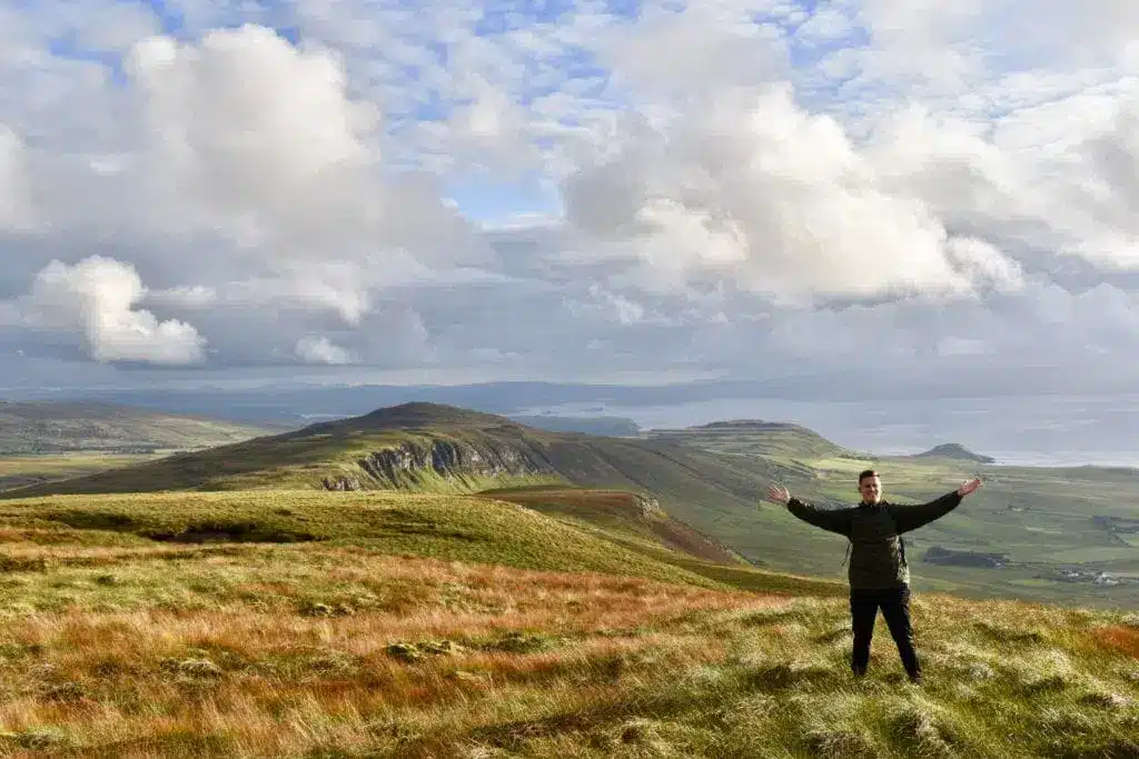 Ein Wanderer mit ausgebreiteten Armen auf einem grasbewachsenen Bergrücken auf Skye
