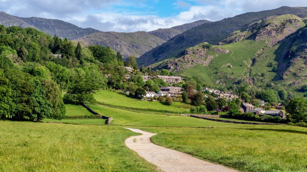 Weg durch eine grüne Wiese zum Dorf Coniston mit Hügeln im Hintergrund