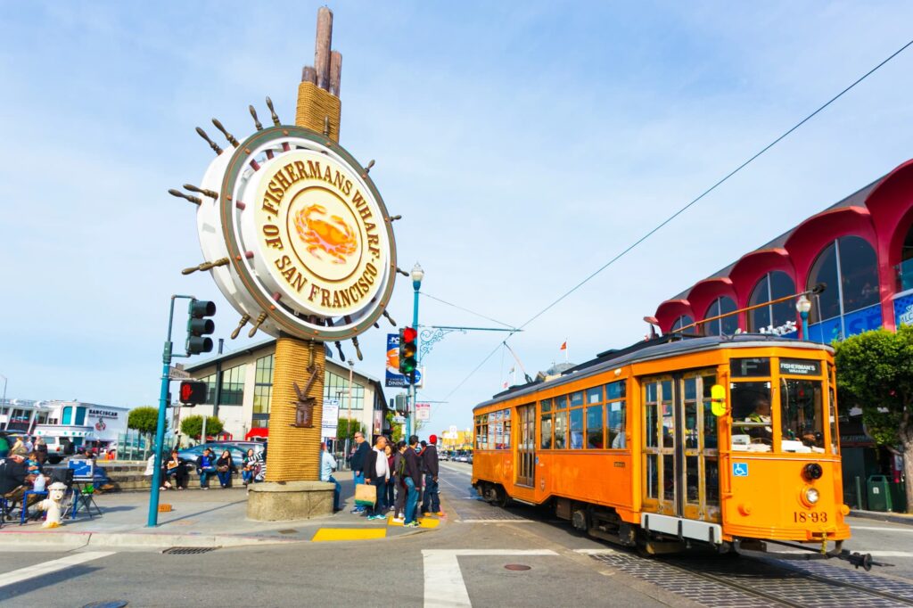 Schild von Fisherman’s Wharf neben einer orangefarbenen Straßenbahn und Gebäuden am Hafen