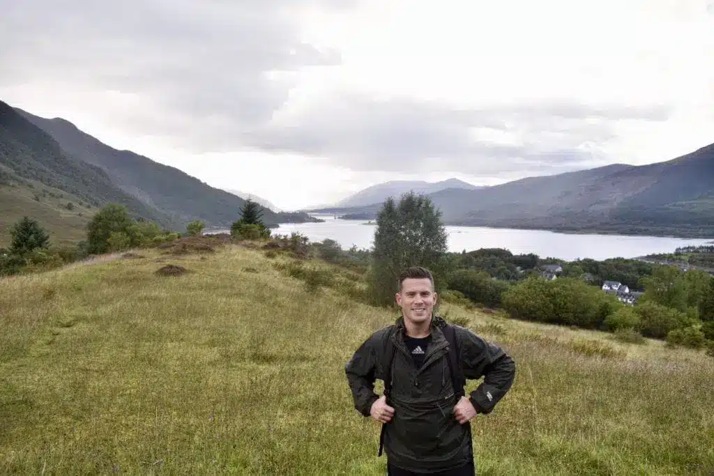 Ein Wanderer auf einem grasbewachsenen Weg mit einem See und Bergen im Hintergrund